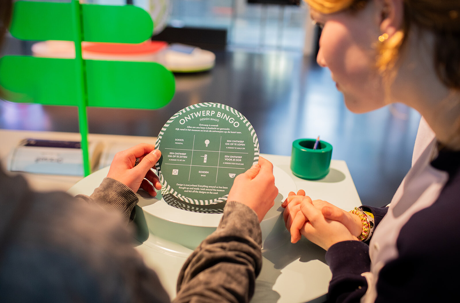 Twee handen houden een 'design bingokaart' vast. Hierop staan opdrachten voor kinderen om ontworpen objecten te vinden, die vervolgens afgekruist kunnen worden.