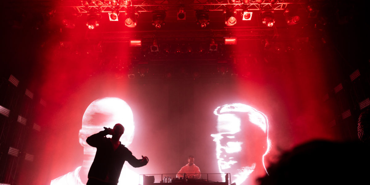 Optreden van The Opposites. De frontman staat op het podium met een microfoon, achter hem op het scherm zijn twee grafische gezichten te zien.