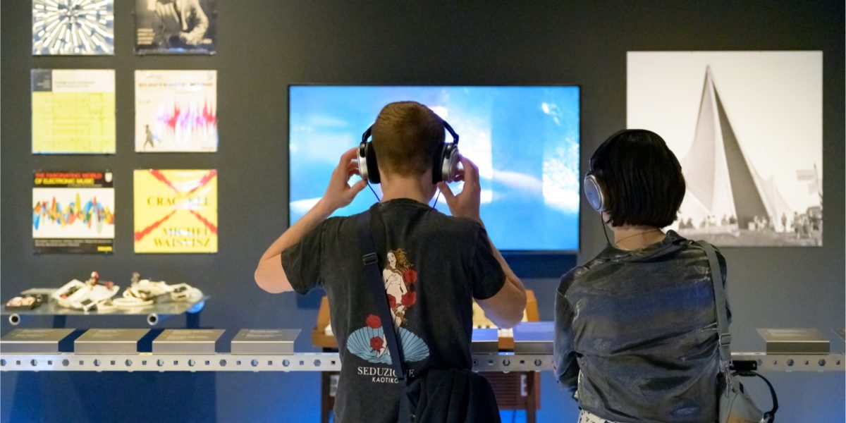 Twee mensen van achter gefotografeerd kijken met een koptelefoon op naar een video op een tv op een muur in een tentoonstelling.