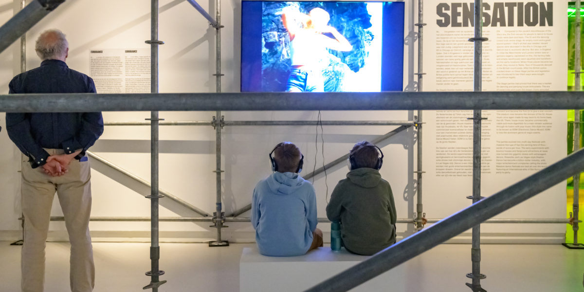 Twee jongens die op een bankje zitten in een tentoonstellingszaal en samen naar een video op een tv op de wand kijken. Ze zijn op de rug gefotografeerd tussen een aantal stijgerbuizen door.