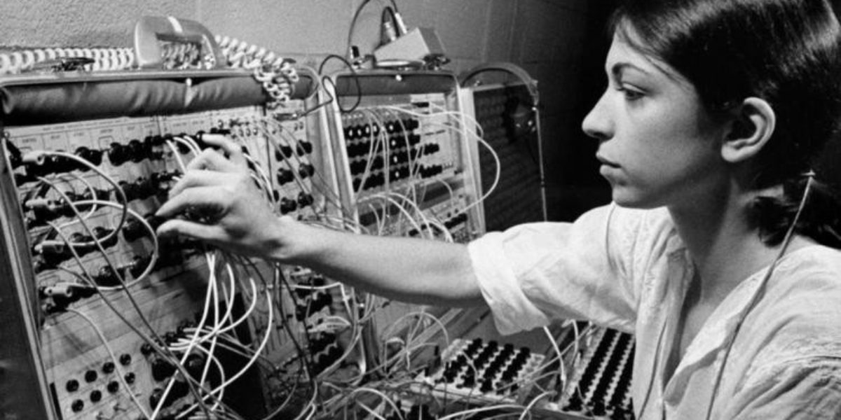 Een oude zwart-wit foto van een vrouw die een analoge synthesizer bediend. De synthesizer ziet eruit als twee opengeklapte koffers vol met draden die ingangen met elkaar verbinden.