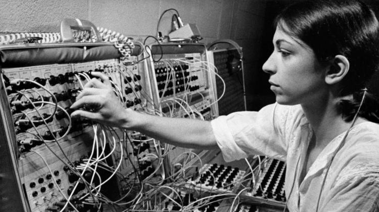 Een oude zwart-wit foto van een vrouw die een analoge synthesizer bediend. De synthesizer ziet eruit als twee opengeklapte koffers vol met draden die ingangen met elkaar verbinden.