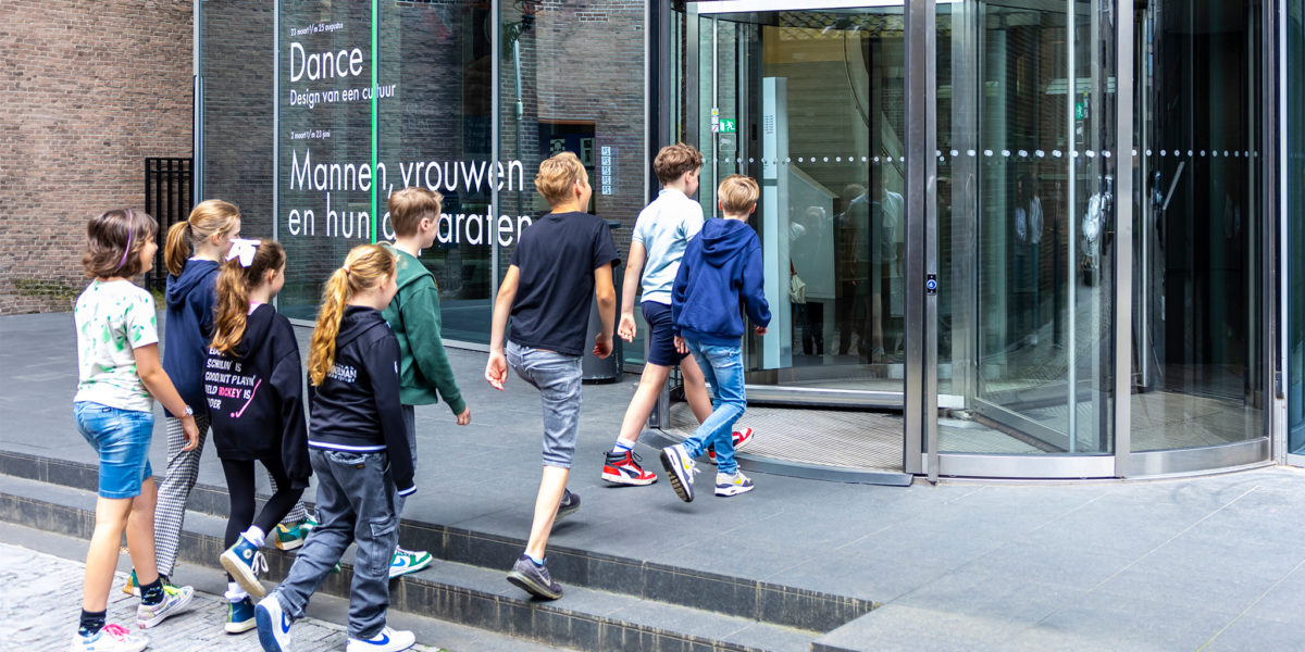 Een klas loopt vanaf de straat via een draaideur Design Museum Den Bosch binnen.