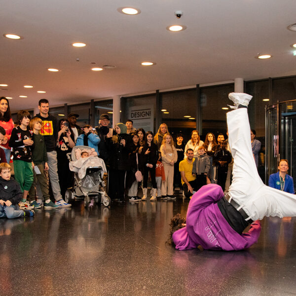 Een foto van een breakdancende jongen die op zijn kop draait, met daaromheen een publiek met veel verschillende mensen.